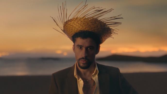 Bad Bunny wearing a spiky straw hat and an open shirt sits on a beach at sunset, looking directly ahead.