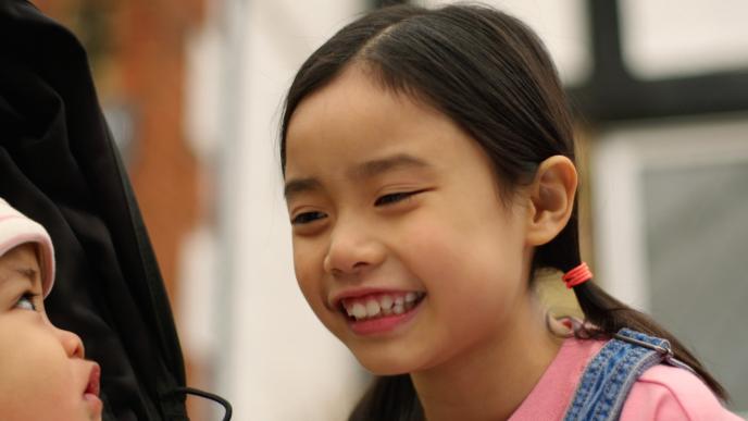 A smiling young girl leans toward a baby in a stroller, sharing a warm, joyful moment outdoors.