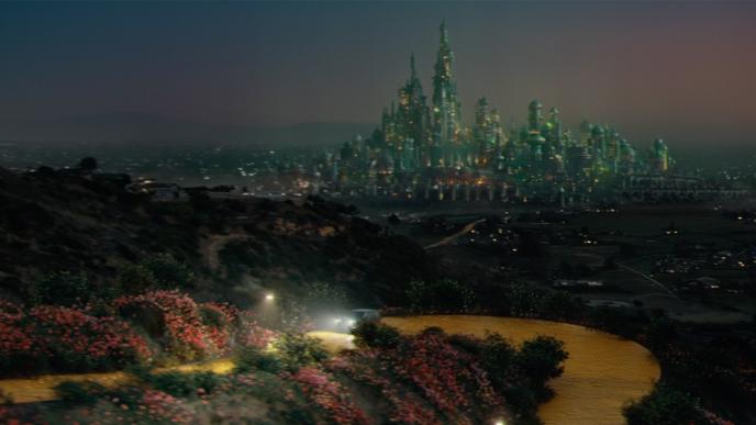 A glowing emerald city rises in the distance as a winding yellow road curves through flowered hills beneath twilight sky.