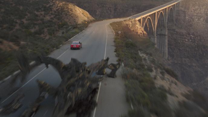 A red car drives down a mountain road at sunset, chased by an alien creature