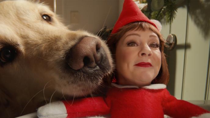 A golden retriever presses its nose toward the camera beside a small elf doll with Melissa McCarthy's comped face in an elf costume