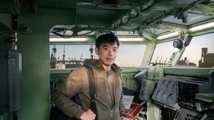 A photograph of an asian man in a ships bridge with a city skyline in the background. 