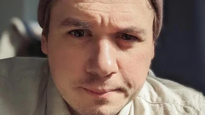 A photograph of a white man wearing a brown beanie hat and a beige shirt looking at the camera.