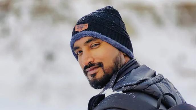 A photograph of an Indian man wearing a puffer jacket and backpack in the snow. 