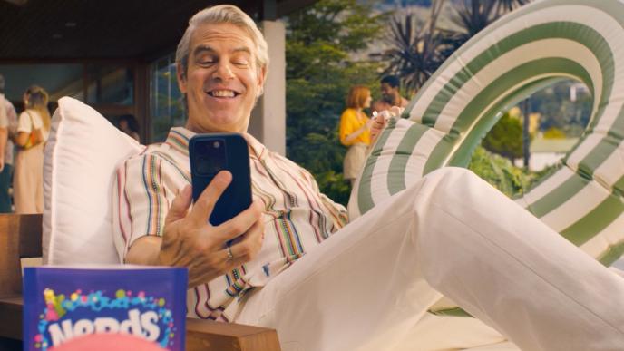 Andy Cohen sits on a pool lounge chair while answering a video call on his cell phone. He is holding an inflatable pool ring