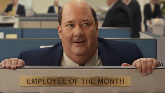 Actor Brian Baumgartner peers over a cubicle wall. There is an "Employee of the Month" placard on the wall