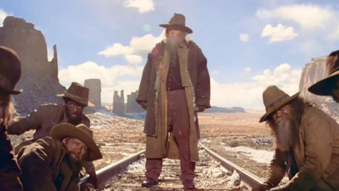 Bearded man stands on railroad tracks in desert, surrounded by crouching men in rugged clothing, under bright sky and rock formations.