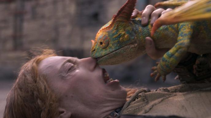 A small CGI dragon sits on a man's chest, biting his nose.
