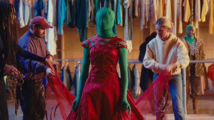 A woman wearing a green screen suit with a red dress over the top there are other people in the background moving and adjusting the fabric.