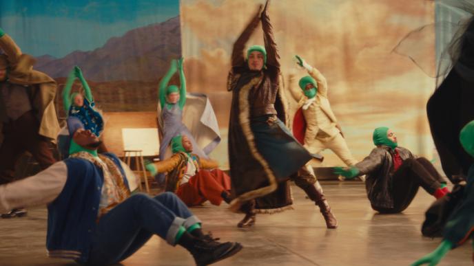 A group of dancers wearing greenscreen suits with TV costumes over the top following a lead dancer in choreography.