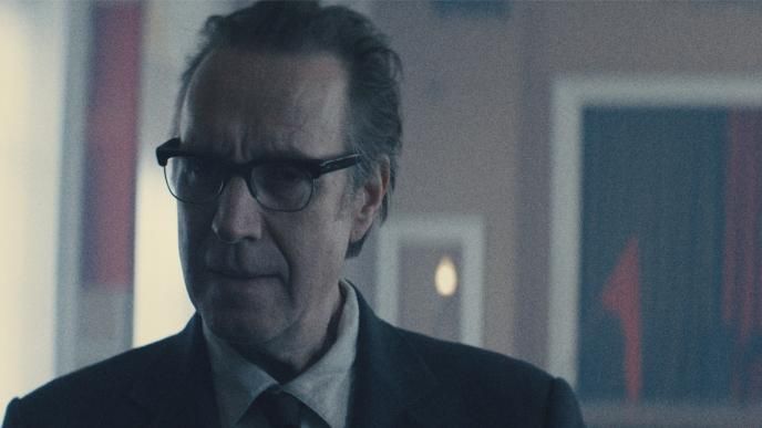 Actor Rhys Ifans in the tv show Star City, wearing glasses and a suit stands indoors, softly lit, with blurred artwork and warm lights in the background.