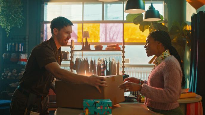 A UPS delivery worker hands a package to a smiling woman inside a colorful boutique with clothing racks and warm lighting.