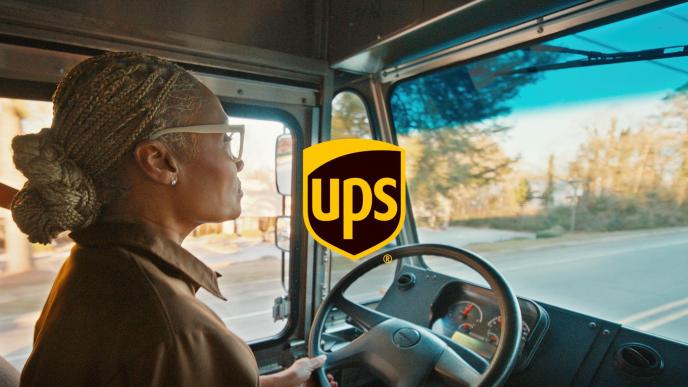 A delivery driver wearing glasses drives a UPS truck, focused on the road, with a large UPS logo overlaid in center.