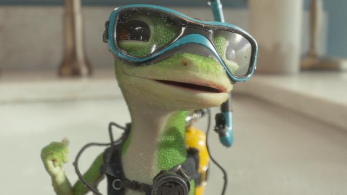 CG animated GEICO gecko wearing blue diving goggles and scuba gear stands in a bathroom sink.