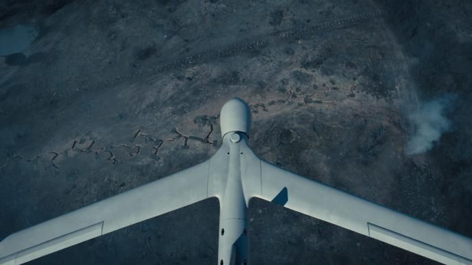 Aerial view of a drone flying over a rugged, war-torn landscape with smoke rising in the distance.