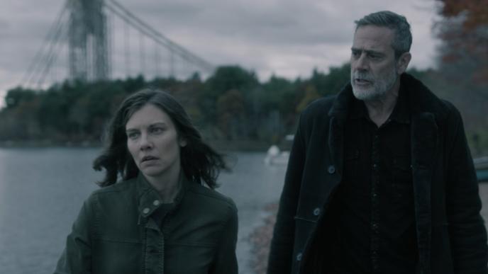 Lauren Cohan as Maggie Rhee and Jeffrey Dean Morgan as Negan Smith, looking somber and walking along a riverside beneath an overcast sky, with a broken suspension bridge and autumn trees behind them.