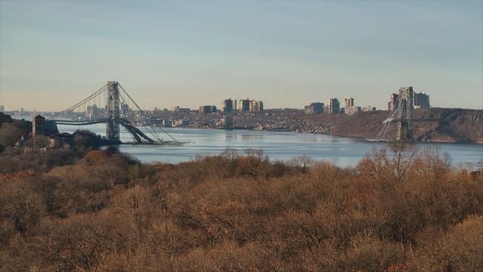 A destroyed George Washington Bridge spans a calm river, surrounded by wooded hills and distant buildings under a clear sky.