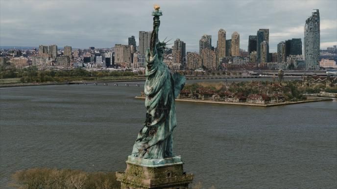 Damaged Statue of Liberty overlooks the river and a decaying city skyline beneath an overcast sky.