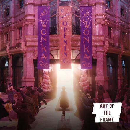 A man walking into an ornate building from which light is spilling onto the street. There are banners hanging from the building that say Wonka and dancers either side of the man.