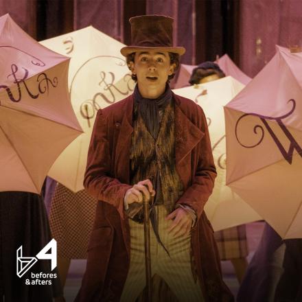 A man standing outside a large ornate building with people behind him dancing with umbrellas that say Wonka on them.