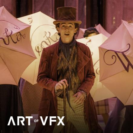 A man standing outside a large ornate building with people behind him dancing with umbrellas that say Wonka on them.