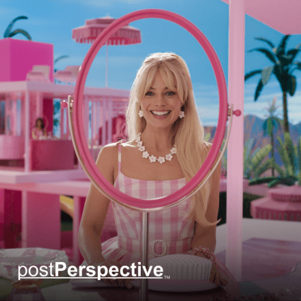 A woman sat looking into the frame of a pink mirror smiling, in the background there are pink houses and a blue sky.