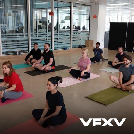 A group of people sat in a glass walled room on yoga matts. 
