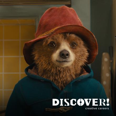 Paddington Bear wearing a red hat and a blue coat stood in a kitchen.