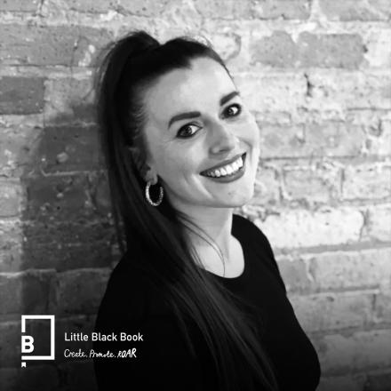 A black and white image of a woman with dark hair smiling against a brick wall.