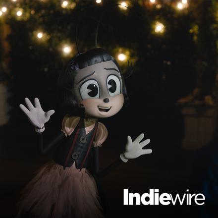 A black and white animated humanoid butterfly stood in a room lit with string lights.