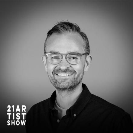 A black and white headshot of a man with the 21 Artists Show logo in the bottom left corner.