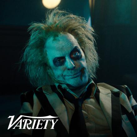 A man wearing black and white facepaint with green fluffy hair is sat looking into the camera.