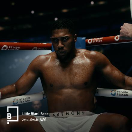 A man wearing boxing shorts and gloves sat in the corner of a boxing ring with a CG crowd behind him.
