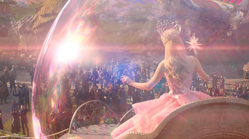 Glinda from Wicked in a CGI bubble floating over Munchkin Land.