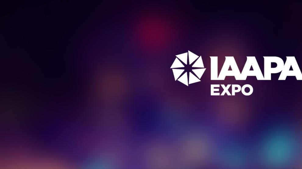 The IAAPA Orlando Expo logo in white on a dark and softly lit background showing multiple colours.