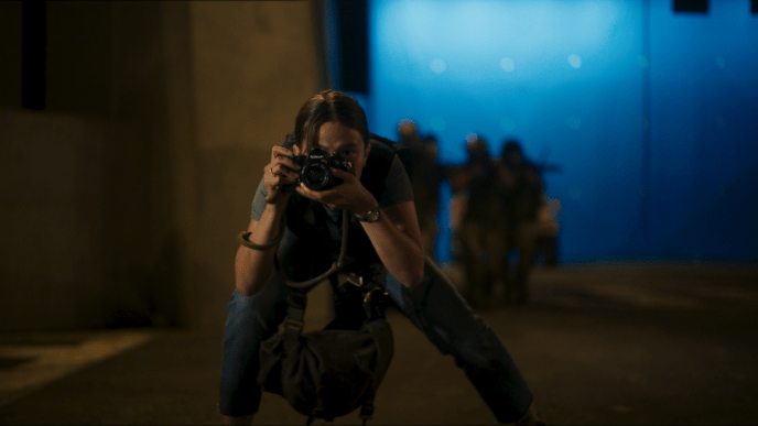 A woman holding a camera and wearing a bulletproof vest is crouching and taking a picture in front of a bluescreen.