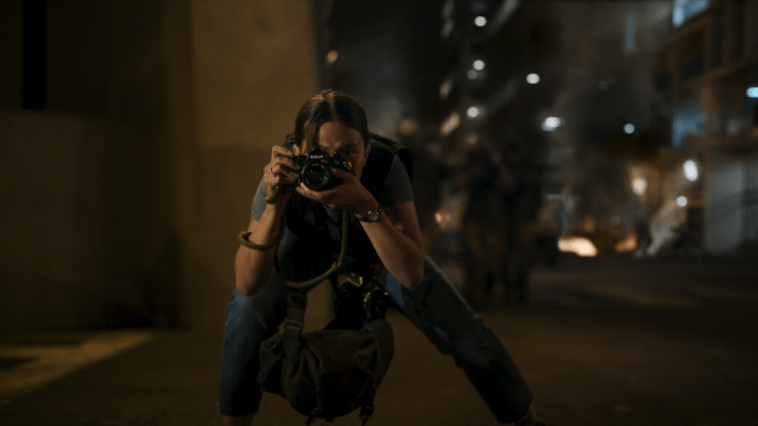 A woman holding a camera and wearing a bulletproof vest is crouching and taking a picture in front of a US street in the dark.