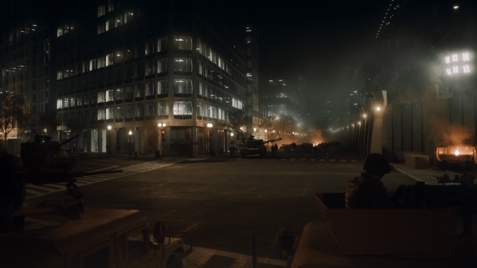 A darkly lit street scene showing fire in the distant background and lights in the high rise buildings.